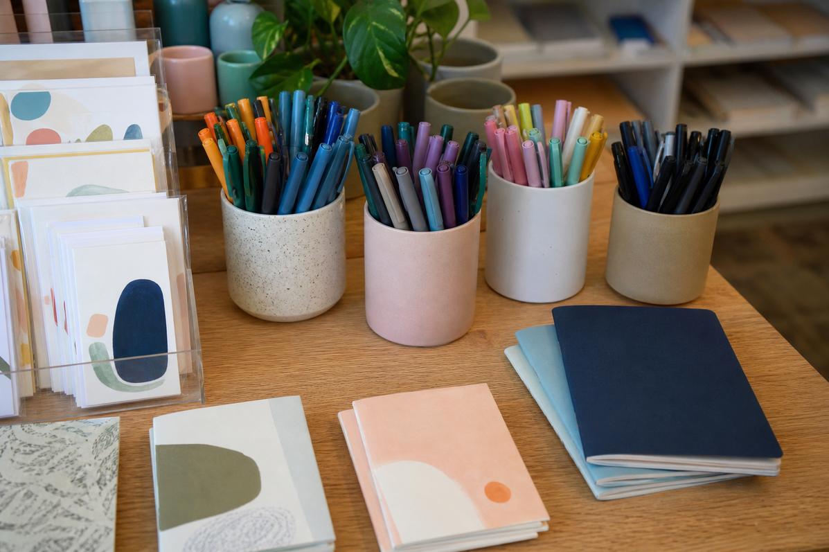 Ceramic cups of coloured pens beside greeting cards and small notebooks